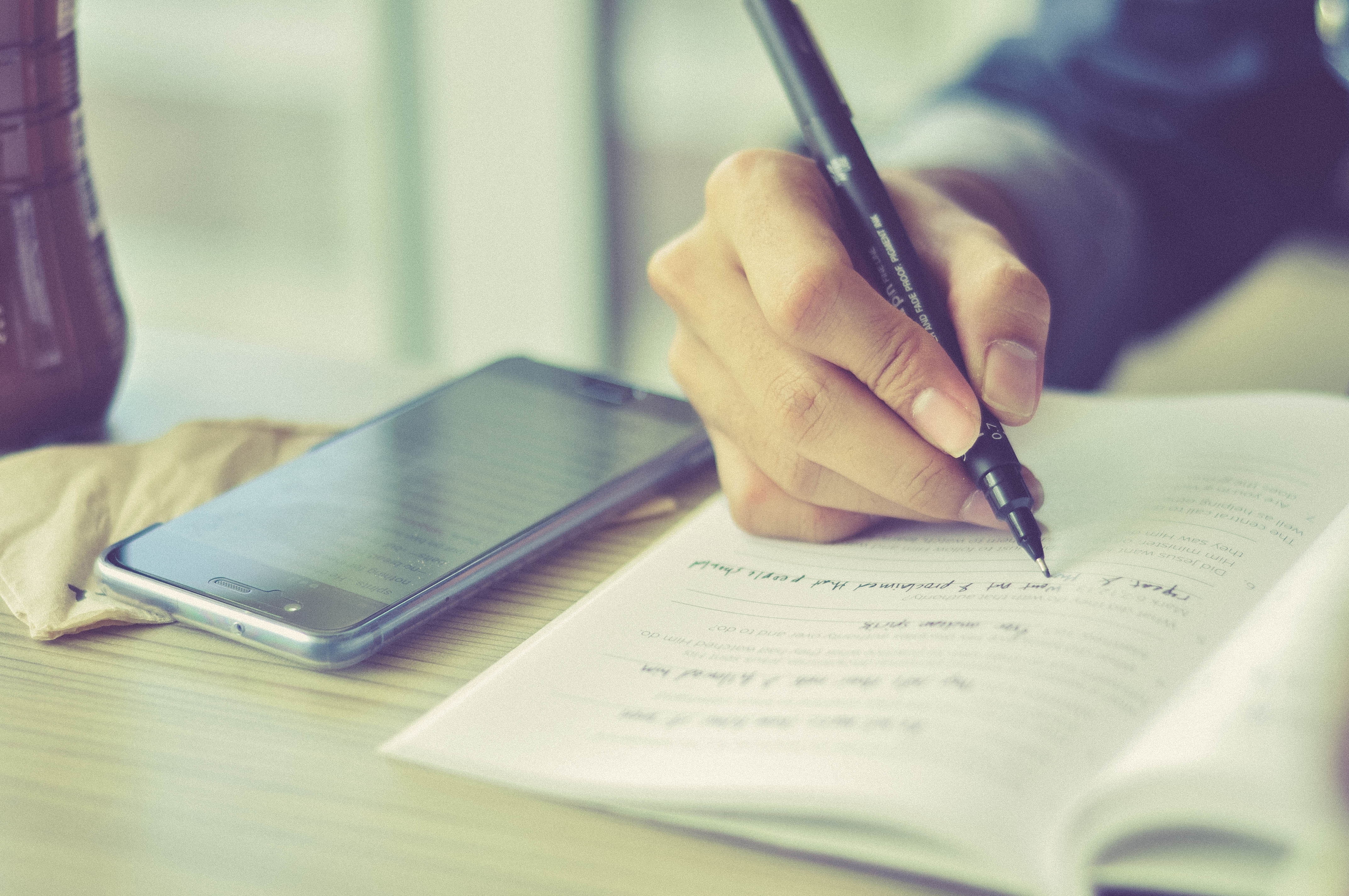 A close-up image of a person writing in a notebook, with a phone laying next to the notebook.